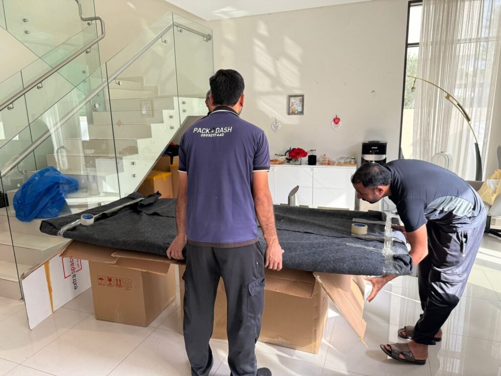 Two PacknDash movers wrapping a dining table in a protective blanket beside a villa staircase in Dubai